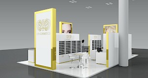 CAD-Visualisierung: Perspektivische Ansicht eines Messestands für die Firma CFB Cosmetics in Weiß mit goldfarbenen Rahmen. Links eine hohe Leuchtstele mit goldenem Rahmen und dem ornamentalen CFB-Cosmetics-Logo, daneben eine Stele mit dem Foto von Augenpartie und Gesicht einer Frau. Dahinter weiße Regale mit vielen Fächern und eine Theke mit CFB-Logo. Ein schwarzer Studioscheinwerfer auf Stativ, weiße Barhocker und ein runder Stehtisch ergänzen den Bereich. Rechts eine graue Rundsäule; der glänzende Boden spiegelt die Aufbauten. CAD-Visualisierung erstellt von Torsten Kraus.