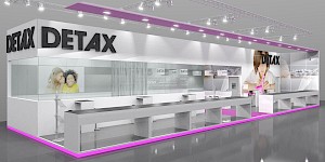 CAD-Visualisierung: Frontalansicht eines Messestands für die Firma Detax, gehalten in Weiß und Grau mit pinkfarbenen Akzenten. Auf der weißen Blende prangt das große schwarze DETAX-Logo. Über dem Stand hängt eine Traverse mit Scheinwerfern, eine pinkfarbene Leuchtlinie zieht sich an der Decke und am Sockel entlang. Links eine durchsichtige Glasvitrine mit dem Foto zweier tuschelnder Kinder, dahinter eine Wand mit einem Wortmuster aus Begriffen wie medical, SILICONES, QUALITY, DETAX und COMPOSITE. Rechts ein großflächiges Foto eines Kindes, das mit pinkfarbenem Silikon modelliert. Lange weiße Theken mit dunklen Deckplatten stehen im Vordergrund, der glänzende Boden spiegelt den Stand. CAD-Visualisierung erstellt von Torsten Kraus.