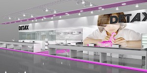 CAD-Visualisierung: Perspektivische Ansicht eines Messestands für die Firma Detax in Weiß und Grau mit pinkfarbenen Akzenten. Über dem Stand hängt eine Traverse mit zahlreichen Scheinwerfern, an der Decke und am Sockel verläuft eine pinkfarbene Leuchtlinie. Links das schwarze DETAX-Logo, dahinter hinter Glas eine Wand mit einem Wortmuster aus Begriffen wie QUALITY, SILICONES und DETAX. Rechts ein großes Foto eines Kindes, das mit pinkfarbenem Silikon modelliert. Lange weiße Theken mit dunklen Deckplatten und offenen Ablagen prägen den Vordergrund, der glänzende Boden spiegelt die Aufbauten. CAD-Visualisierung erstellt von Torsten Kraus.