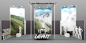 CAD-Visualisierung: Frontalansicht eines Messestands für die Firma Leatt in Weiß und Grau mit Naturmotiven. Hinter einem durchscheinenden Vorhang aus dünnen Stäben zeigt eine hinterleuchtete Wand das LEATT-Logo vor einem Foto grüner Berglandschaft mit Wolken; oben werfen mehrere Spots Licht auf die Fläche. Links und rechts zwei hohe Bildstelen mit weiterer Berglandschaft. Auf einem Tisch in der Mitte drei Helme auf Ständern, davor das schwarz-weiße LEATT-Blocklogo. Links ein Warenträger mit gelben Nackenschützern auf weißen Büsten, rechts mehrere weiße Schaufensterfiguren. CAD-Visualisierung erstellt von Torsten Kraus.