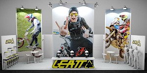 CAD-Visualisierung: Frontalansicht eines Messestands für die Firma Leatt in Weiß und Gelb mit Grau. Drei große hochformatige Leuchttafeln zeigen Actionfotos eines BMX-Fahrers, eines Mannes auf einem Motorrad mit erhobener Hand und eines Motocross-Fahrers; Spots werfen deutlich sichtbares Licht auf die Motive. Links an der Wand das gelbe LEATT-Logo und ein Warenträger mit gelben Nackenschützern auf weißen Büsten. In der Mitte eine weiße Theke mit gelbem LEATT-Logo und drei Helmen. Dazu weiße Stehtische mit Barhockern, rechts graue Schaufensterfiguren. Der Boden ist gefliest. CAD-Visualisierung erstellt von Torsten Kraus.