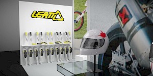 CAD-Visualisierung: Nahansicht eines Ausschnitts eines Messestands für die Firma Leatt. An einer weißen Wand hängt das gelbe LEATT-Logo, darunter präsentieren zwei Reihen weißer Büsten gelb-schwarze Nackenschützer. Im Vordergrund ein durchsichtiger Glastisch mit einem Helm auf einem Ständer, der einen roten Farbspritzer trägt. Im Hintergrund Fotos eines Motorrad-Auspuffs und eines BMX-Rads. CAD-Visualisierung erstellt von Torsten Kraus.